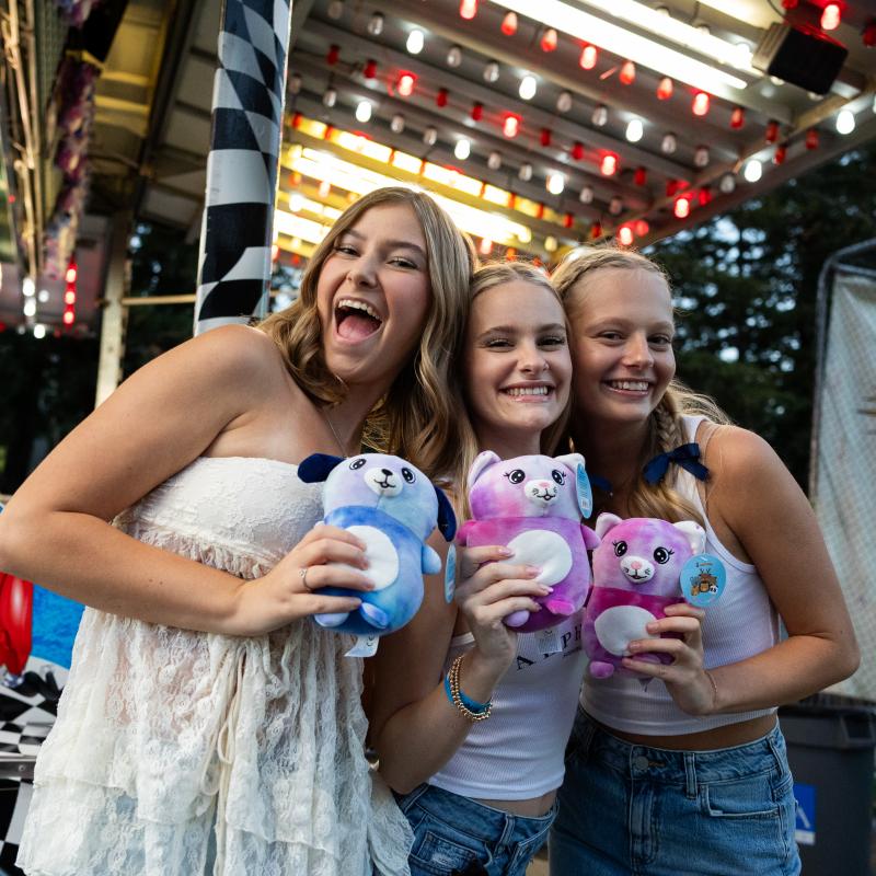 Three students holding stuffed animals in front of a carnival game.