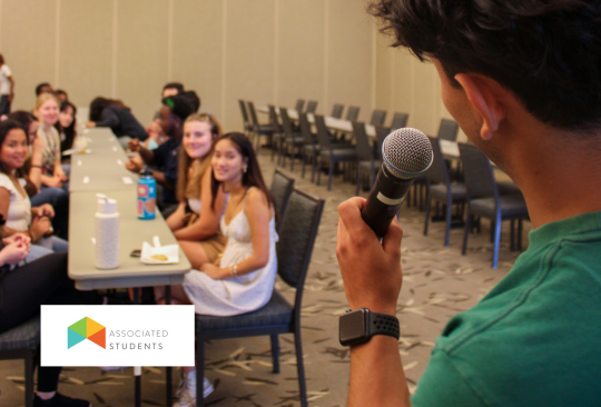 Student speaking into a microphone facing towards rows of seated students.